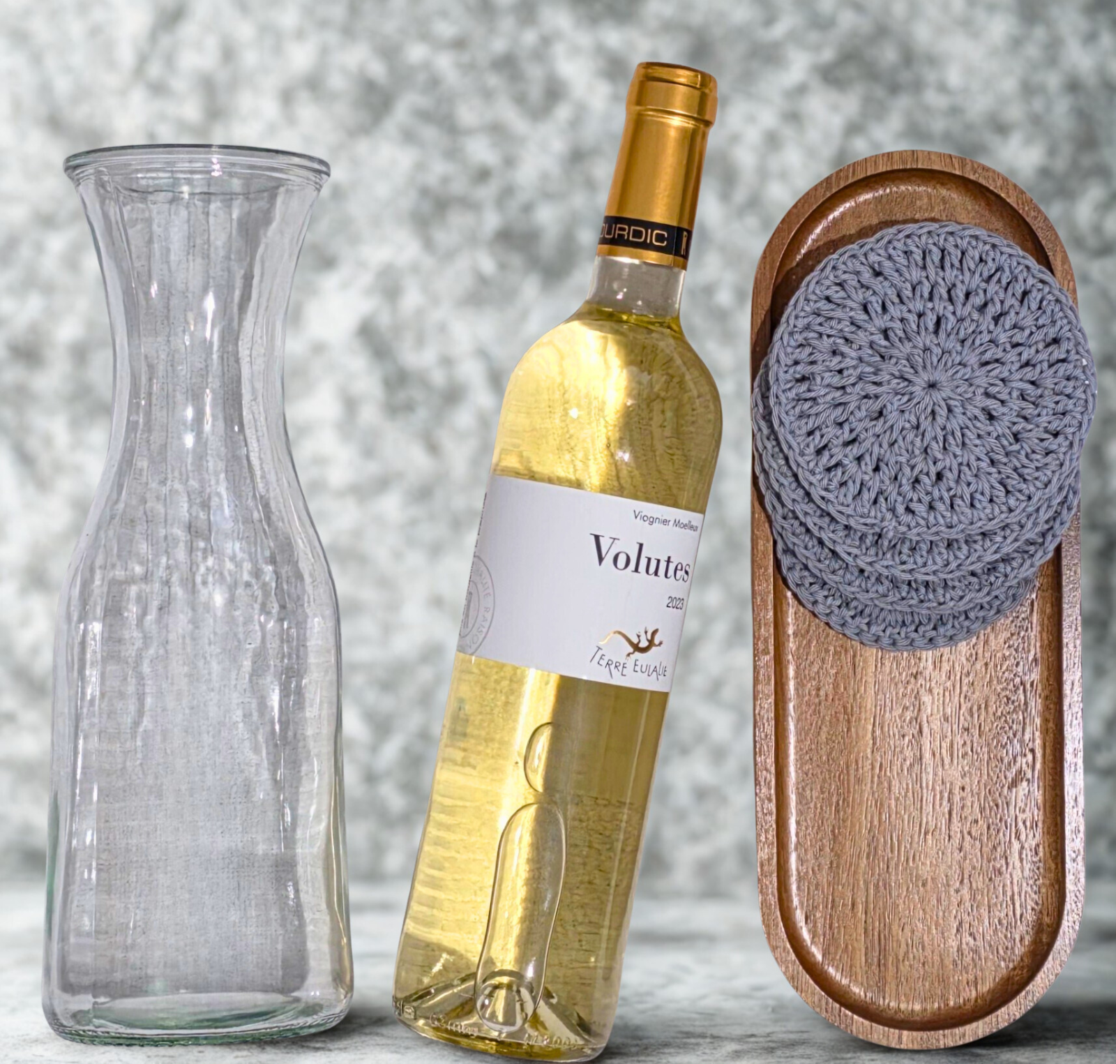 Wine bottle labeled 'Volutae' with a glass carafe and wooden tray with crochet coasters on a neutral background
